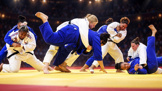 Several female judoka in white and blue judogi compete on a yellow tatami under arena lights, with athletes mid-throw and on the ground.