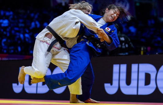 A judoka in a blue gi and a judoka in a white gi grip each other during a standing exchange on the tatami.