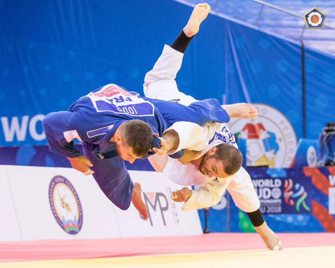 A judoka in a blue judogi throws an opponent in a white judogi during a match on a yellow tatami.