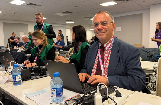 Man wearing glasses and a red lanyard works on a laptop at a desk in a busy media room, with water bottles, cables and other journalists nearby.