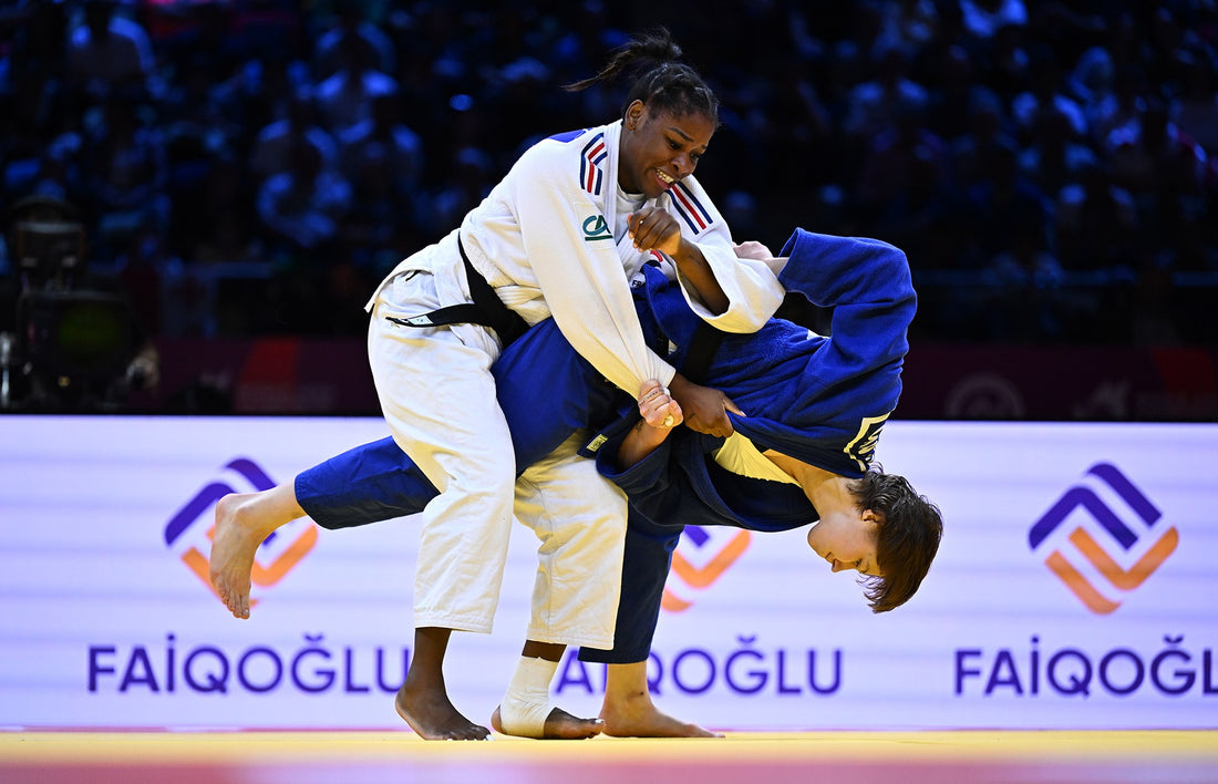 A judoka in a white judogi lifts an opponent in blue during a throw, with a blue-lit crowd in the background.