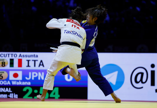 Two judoka in white and blue judogi compete on a mat, with a scoreboard visible behind them.