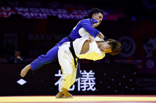 A judoka in a blue gi throws an opponent in a white gi during a judo contest on a brightly lit mat.