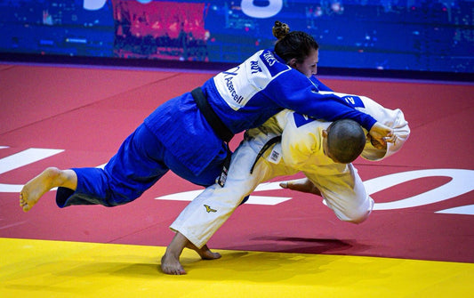 A judoka in a blue gi drives an opponent in a white and yellow gi toward the tatami during a contest.