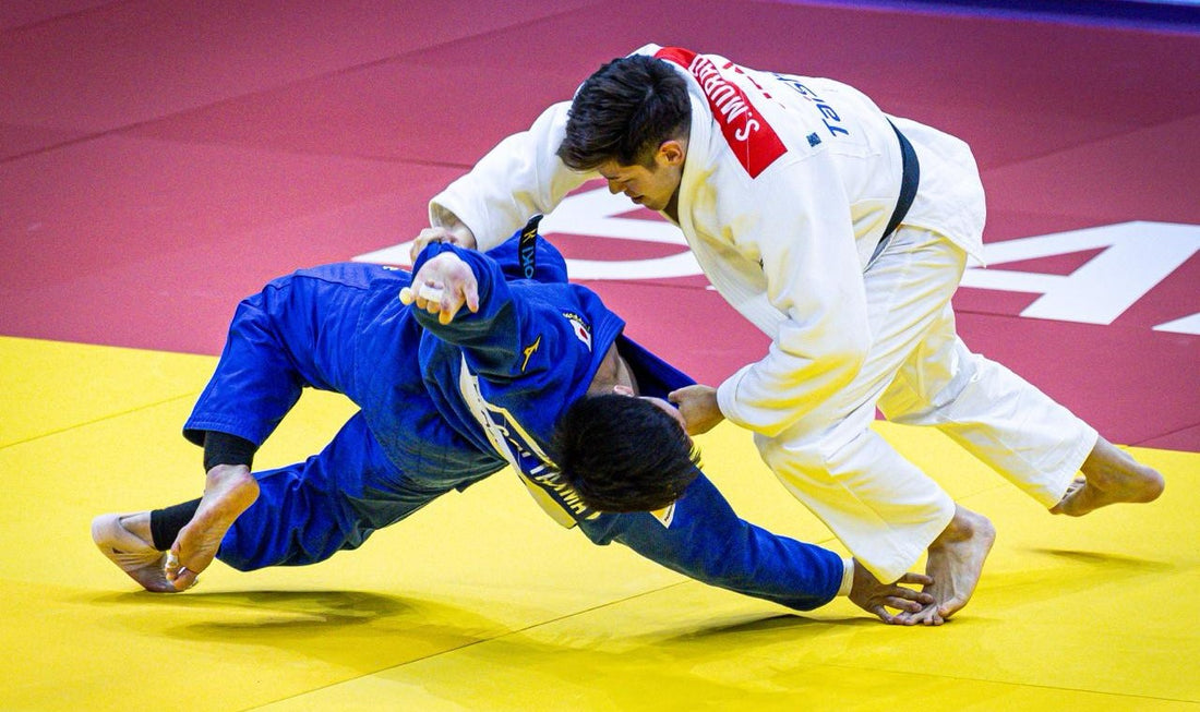 A judoka in a white gi controls an opponent in a blue gi during a throw attempt on a yellow and red tatami.