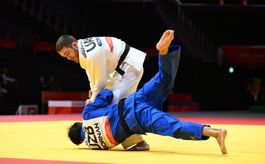 A judoka in a white judogi controls an opponent in a blue judogi during a throw on a yellow mat.