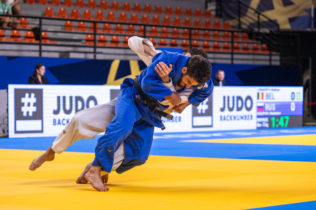 Two judoka in blue and white grapple on a yellow tatami, with orange arena seats behind them.