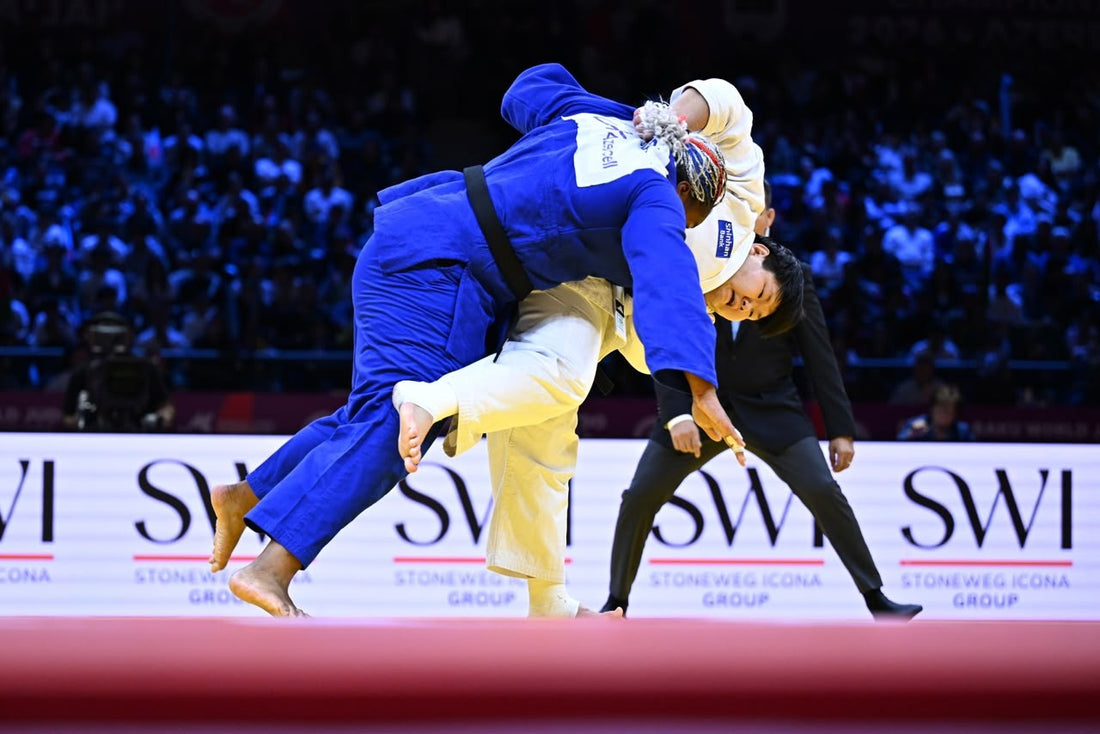 A judoka in white lifts a judoka in blue during a throw, while a referee watches on the competition mat.