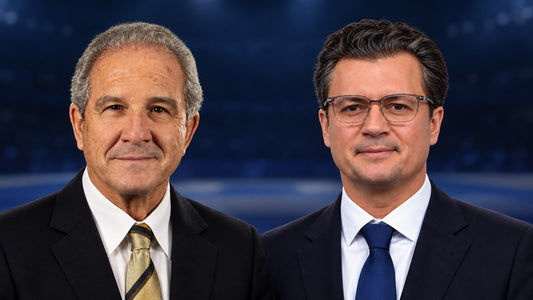 Two men in dark suits, white shirts and ties pose side by side against a blurred blue background.