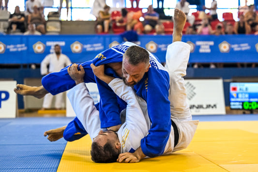 A competitor in a blue judogi grapples with an athlete in a white judogi on a yellow and blue tatami, with spectators in the background.