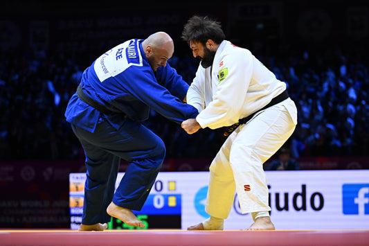 A judoka in a blue judogi and a judoka in a white judogi grip each other on a competition mat.