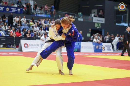 Two female judoka in blue and white judogi grip each other during a contest on a yellow tatami, with spectators behind them.