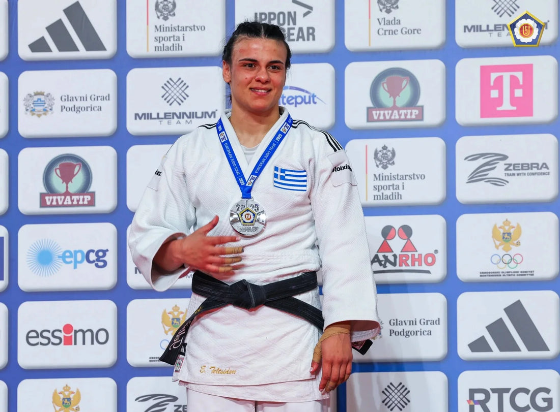 A judoka in a white judogi and black belt wears a medal and Greek flag patch while standing before a sponsor backdrop.