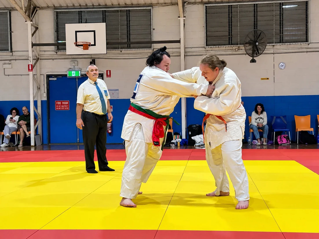 Two people in white Judo uniforms grip each other on a yellow tatami, with an official and seated spectators nearby.