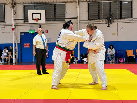 Two people in white Judo uniforms grip each other on a yellow tatami, with an official and seated spectators nearby.