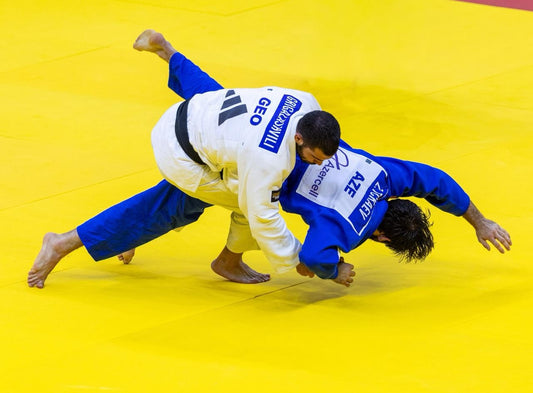 A judoka in a white gi throws or controls a judoka in a blue gi on a bright yellow tatami.