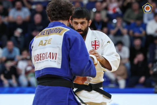 Two judoka in blue and white judogi face each other on the tatami, with spectators blurred in the background.
