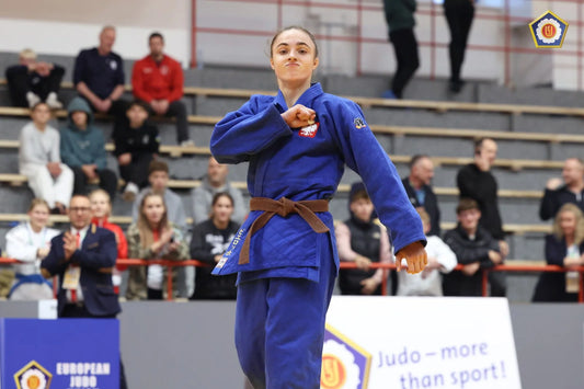 A judoka in a blue judogi and brown belt stands on the mat with one arm raised, with spectators behind her.