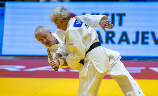 Two judoka in white judogi and black belts practise a standing technique on a yellow and red mat.
