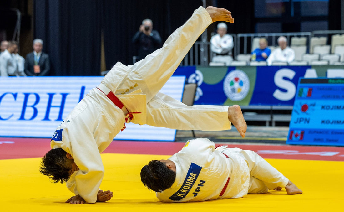 Two judoka in white judogi on a yellow mat, with one athlete lifted upside down while the other lies on the mat.