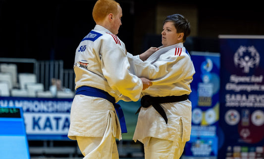 Two judoka in white judogi grip each other on a tatami, with a blue competition display and event signage in the background.