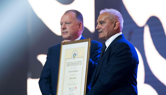Two men in dark suits stand on a stage, with one holding a large framed certificate.