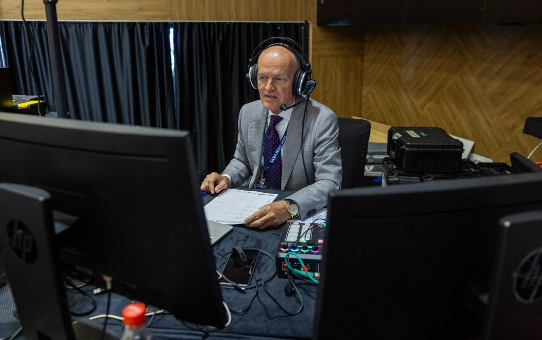 A man wearing a headset and grey suit sits at a commentary desk surrounded by monitors, cables and audio equipment.