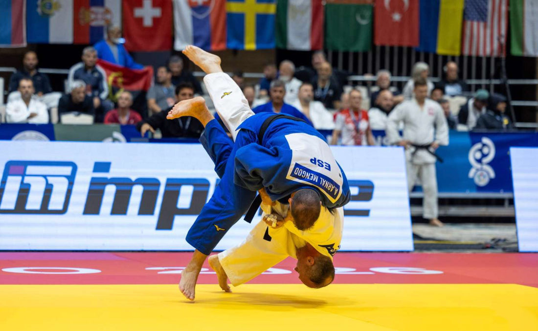 A judoka in blue throws an opponent in yellow on a competition tatami, with spectators and national flags behind them.