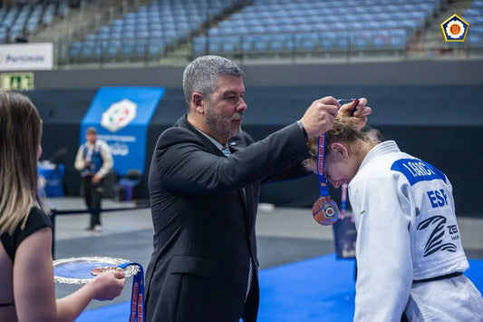 A man in a dark suit places a medal over the head of a judoka in a white judogi on a blue mat.